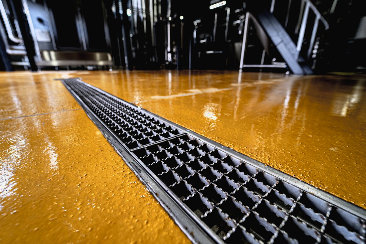A stainless steel trench drain set across a commercial kitchen floor, the floor wet around it, kitchen equipment in the background