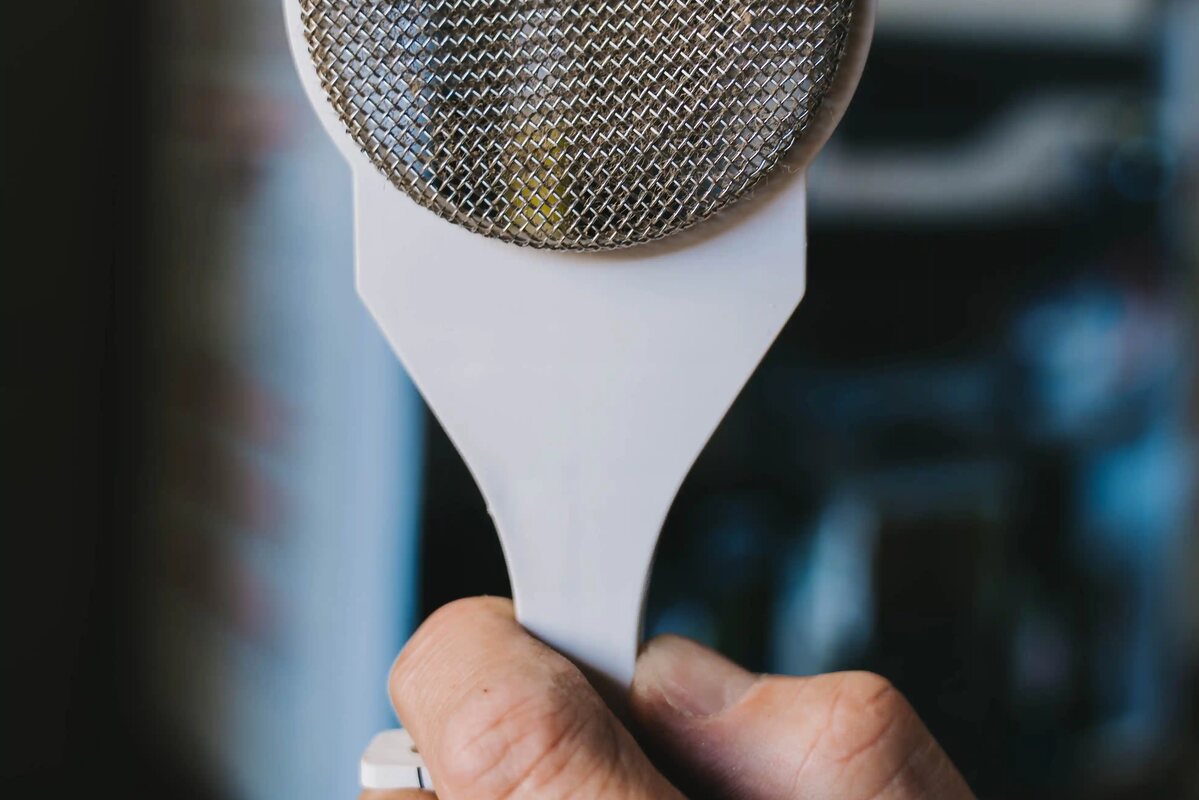 A shower head held up in one hand, the mesh face toward the camera, blurred pipework behind