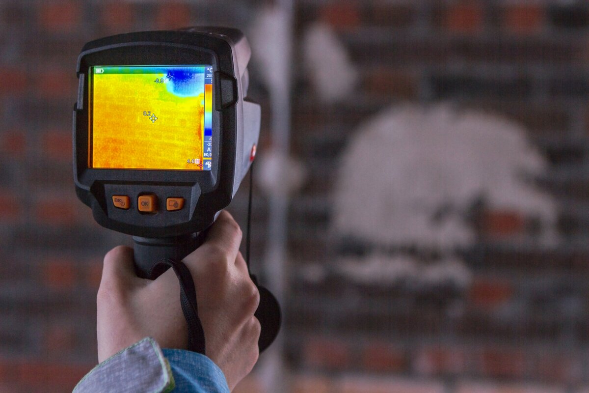 A hand holding a thermal imaging camera against a wall, its screen showing the temperature of the plaster